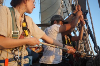 Members of A Watch set the jib as the sun rises. Stella Klema (Smith College) tails the line off the pin, while Kevin Ferreira (SUNY Maritime) and Emily Volkmann (Smith College) work together to sweat the halyard.
