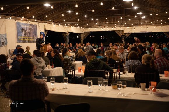 Williams-Mystic alums sit at a large dinner event while Executive Director Tom Van Winkle speaks at a podium